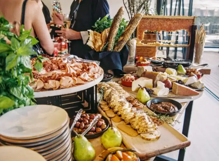 Ein HOMCOM-Tisch mit verschiedenen Käsesorten, Brot, Obst, Wurstwaren und Dips; im Hintergrund wird geplaudert.
