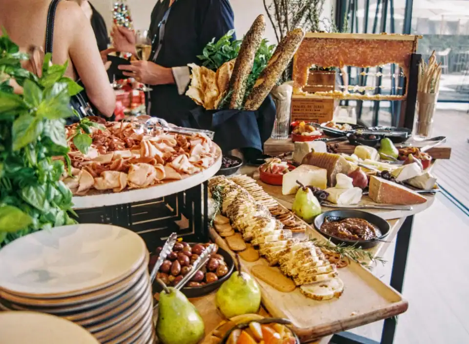 Ein HOMCOM-Tisch mit verschiedenen Käsesorten, Brot, Obst, Wurstwaren und Dips; im Hintergrund wird geplaudert.