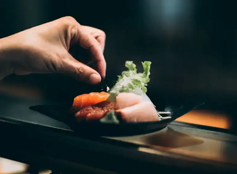 Eine Hand legt Garnierungen auf einen Teller mit verschiedenen Sashimi und Blattgemüse auf einem HOMCOM-Tisch.