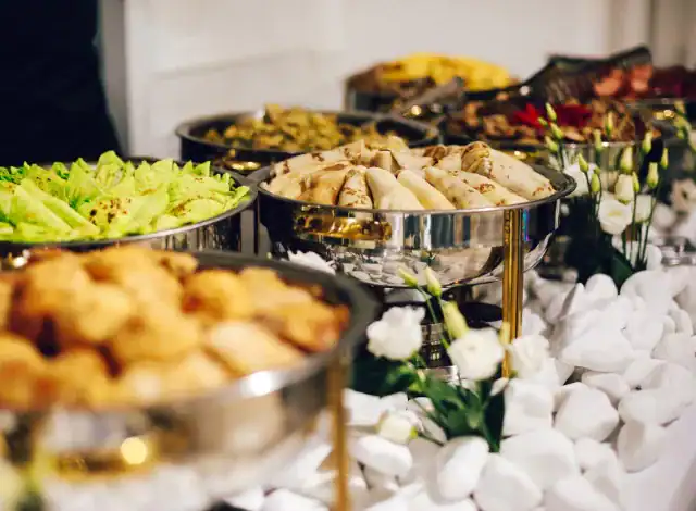 Ein HOMCOM-Buffet-Tisch mit warmen Gerichten und Salaten in Tabletts, umgeben von weißen Dekosteinen und Blumen.