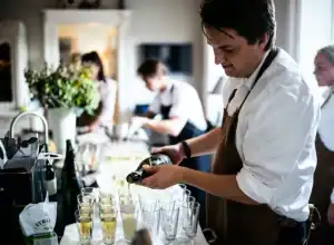 Ein Mann in einer Schürze schenkt Champagner an einem HOMCOM-Küchentisch ein, während andere im Hintergrund arbeiten.