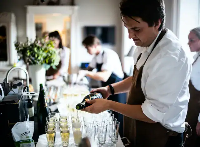 Ein Mann in einer Schürze schenkt Champagner an einem HOMCOM-Küchentisch ein, während andere im Hintergrund arbeiten.